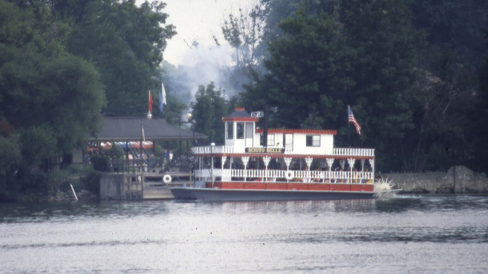 A red and white sternwheeler paddle-wheeler under way on the river past a riverside landing